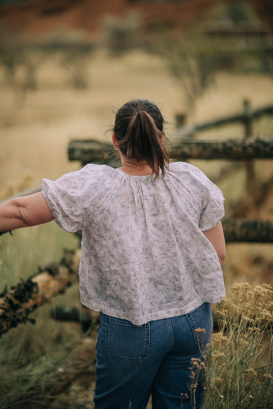 Female model stands in field wearing a button down, floral print, short sleeve, blouse and medium wash jeans.